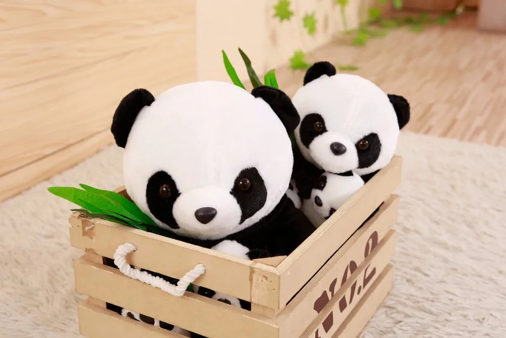 Two panda plush toys in a wooden crate with bamboo leaves on a soft surface.